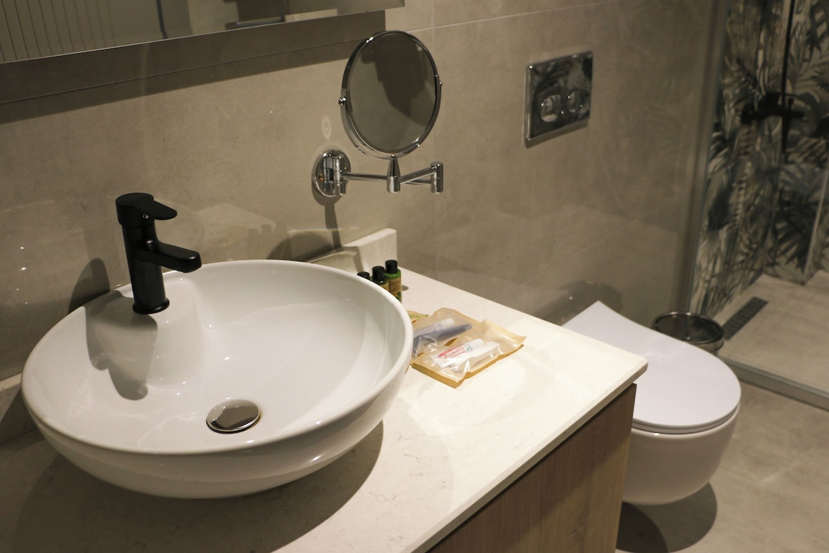 A bathroom with a white sink, black faucet, and a mirror above it. There is a toilet and a shower visible in the background.