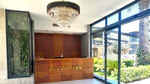 A reception area with a modern design, featuring a wooden reception desk, a chandelier, and large windows offering a view of the outdoors. -  ARCADIA OF BODRUM (Bodrum)