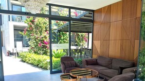 A modern indoor seating area with a glass wall, a wooden geometric wall panel, and a view of outdoor plants and flowers. -  ARCADIA OF BODRUM (Bodrum)