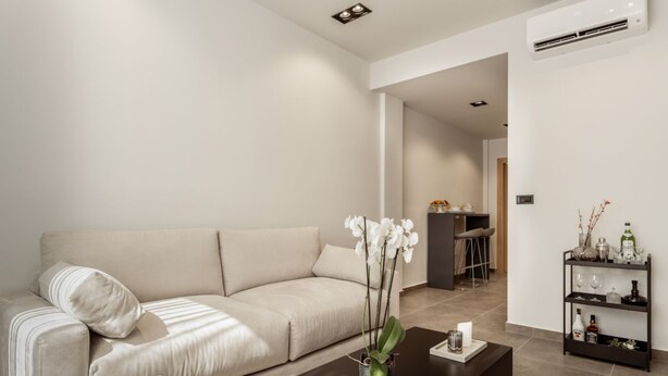 A modern living room with a sofa, a coffee table, and a small plant. There is a bar area in the background with stools and a shelf with bottles and glasses. - La Maison Suite   Suite 2 (Zakynthos)