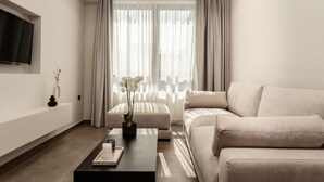 A modern living room with a beige sofa, a coffee table with a vase and candle, a TV on the wall, and sheer curtains. - La Maison Suite   Suite 2 (Zakynthos)