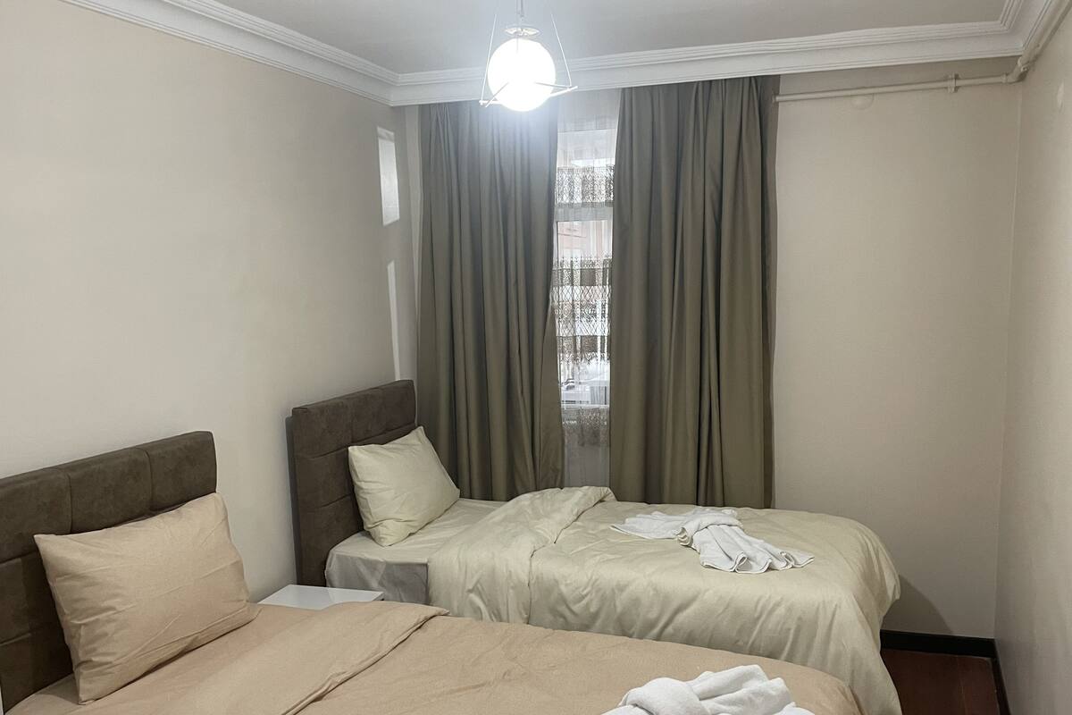A hotel room with two beds, beige bedding, a headboard, a nightstand, and a window with curtains.
