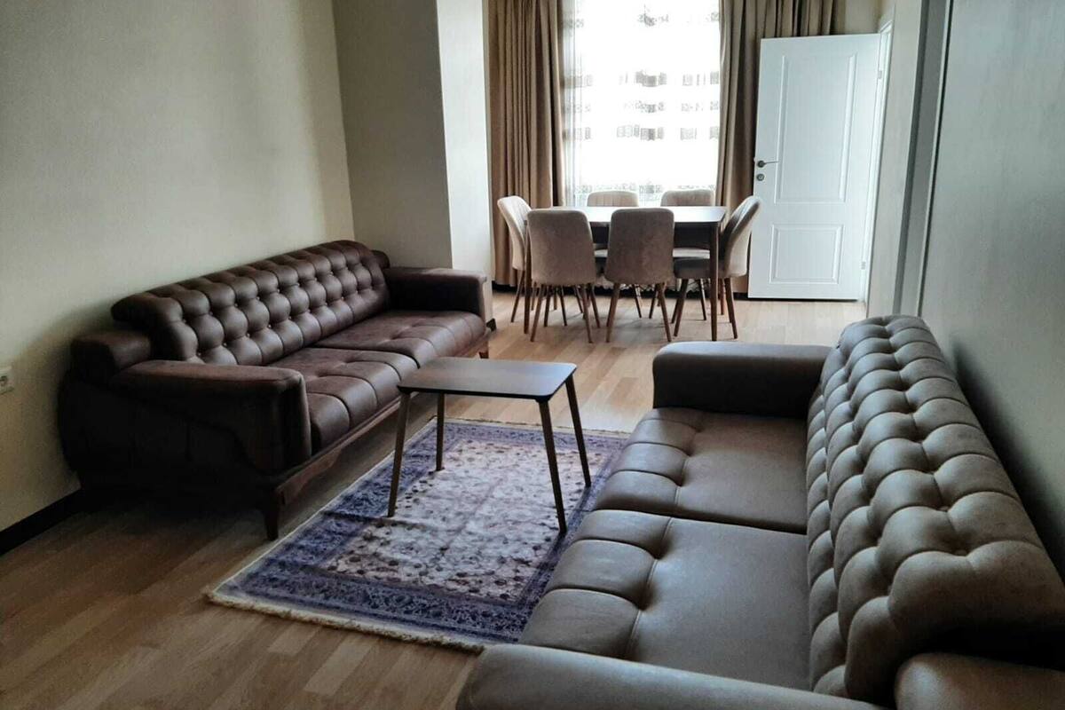 A living room with a brown leather sofa, a matching ottoman, a small coffee table, and a patterned rug. There is a dining area with a table and chairs in the background.