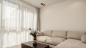 Ett modernt vardagsrum med en beige soffa, ett mörkt soffbord och tunna, genomskinliga gardiner.