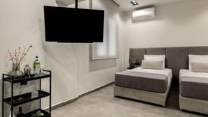 A modern hotel room with a large flat-screen TV mounted on the wall, a bed with grey bedding, a bedside table with a lamp, and a small shelf with decorative items. - La Maison Suites   Studio 6 (Zakynthos)
