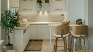Ett modernt kök med vita skåp, en köksö, två barstolar, en krukväxt och en skål med frukt på bänkskivan.