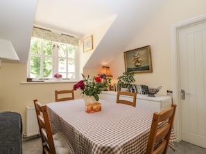 A dining area with a checkered tablecloth, wooden chairs, a vase of flowers, a radiator, a window with a view of greenery, a bookshelf, and a door.