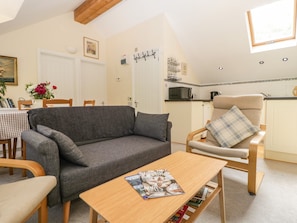 A cozy living room with a grey sofa, a wooden coffee table with magazines, a dining area with a checkered tablecloth, and a kitchen with a microwave and kettle.