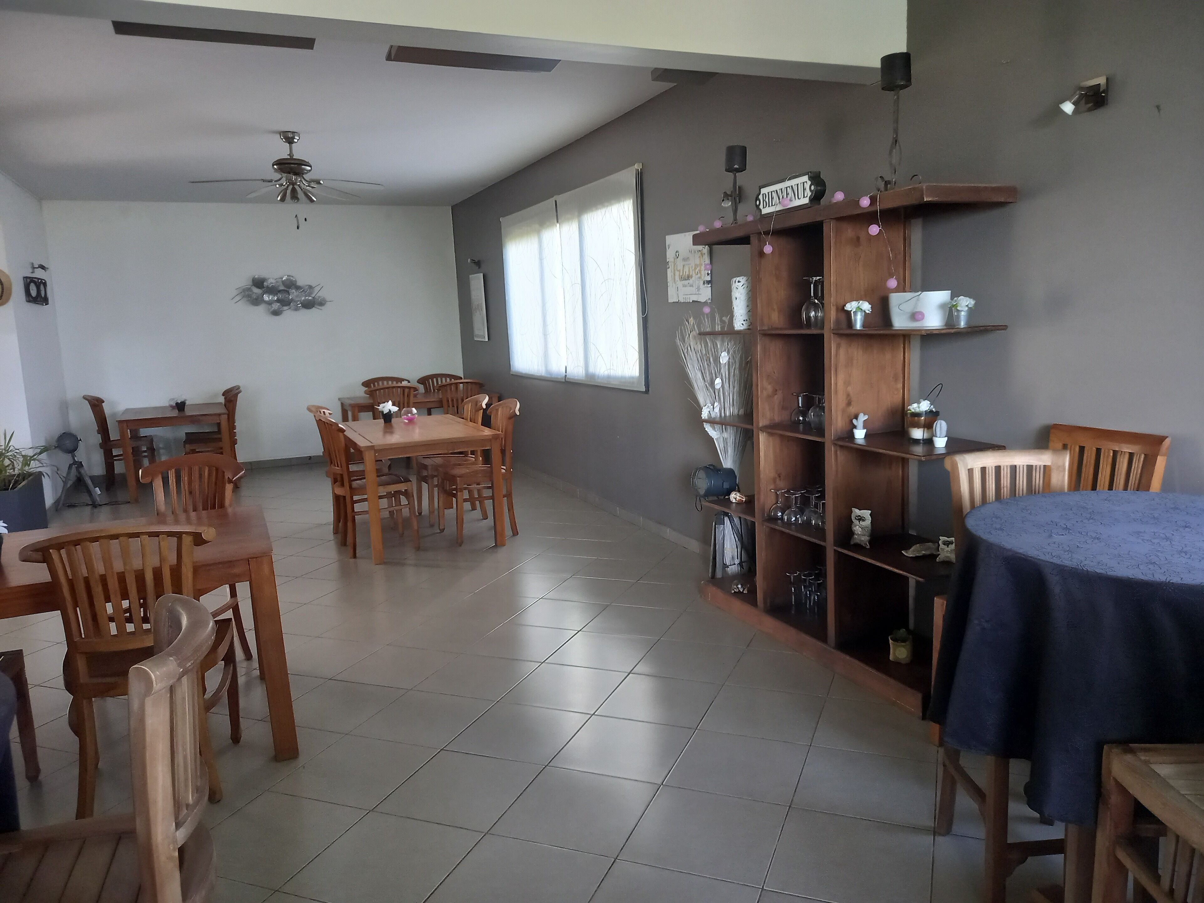 Uma área de refeições com mesas e cadeiras de madeira, um ventilador de teto e uma estante decorativa com diversos objetos.