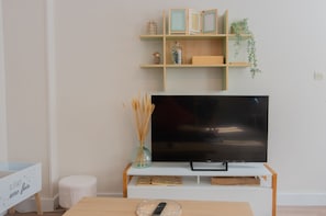 A living room with a flat-screen TV on a white stand, wooden shelves above, and a small table with a basket. - Bergerie du Centre-ville (Dieppe)