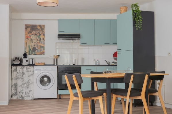 A compact kitchen with teal cabinets, a washing machine, and a dining area with wooden chairs. - Bergerie du Centre-ville (Dieppe)