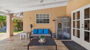 A covered outdoor area with a ceiling fan, a sofa with blue cushions, a refrigerator, and a small table with a plant.