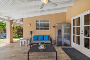 A covered outdoor area with a ceiling fan, a sofa with blue cushions, a refrigerator, and a small table with a plant. - Private Pool Oasis 4BR Home Near Palm Beach, Aruba (Noord)