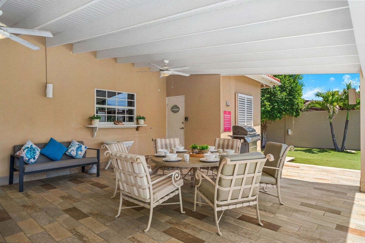 Une terrasse extérieure avec une table ronde, des chaises en osier blanc, un ventilateur de plafond et un barbecue.