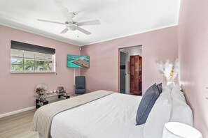Une chambre à coucher avec un lit, un ventilateur de plafond, une fenêtre avec des stores, un bureau avec une chaise et un tableau accroché au mur.