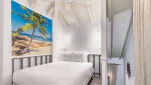 Une chambre à coucher avec un lit, un tableau représentant une scène de plage, un ventilateur de plafond et une fenêtre avec des rideaux.