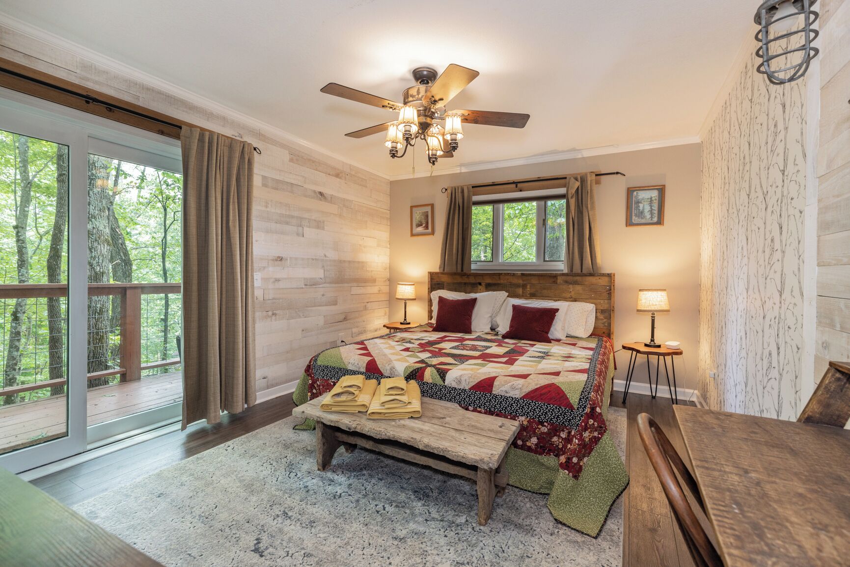 A bedroom with a wooden headboard, a large bed with a quilt, two bedside lamps, a wooden bench, and a view of trees through a sliding glass door.