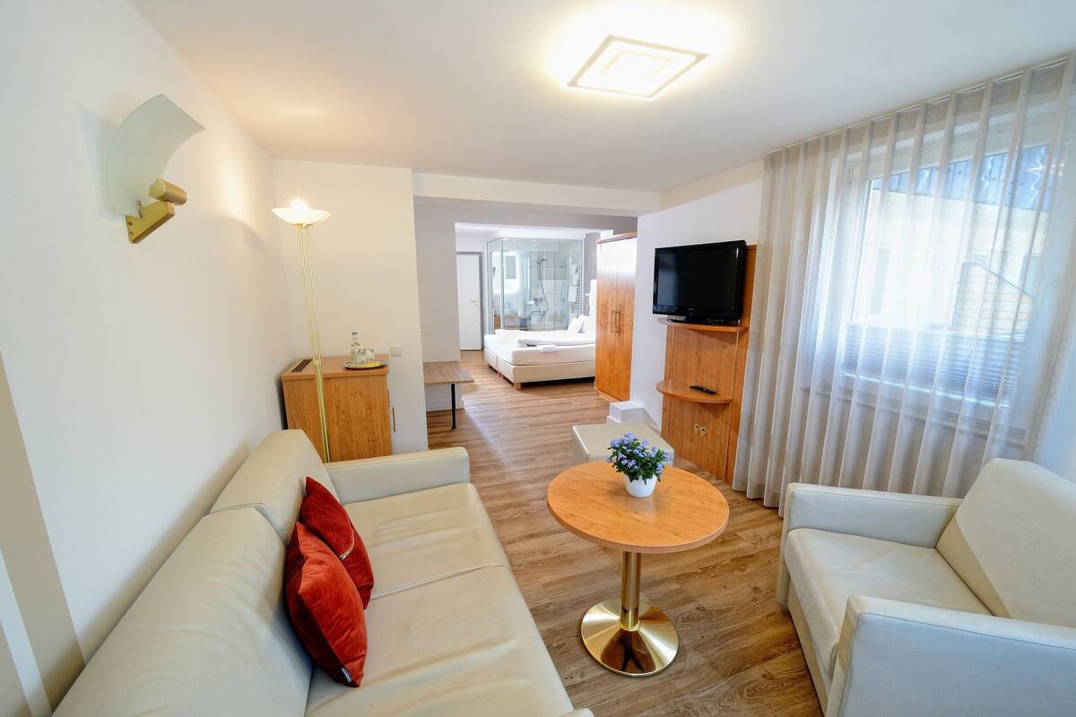 A modern hotel room with a sofa, a round table with a plant, a TV, and a view of a bedroom.