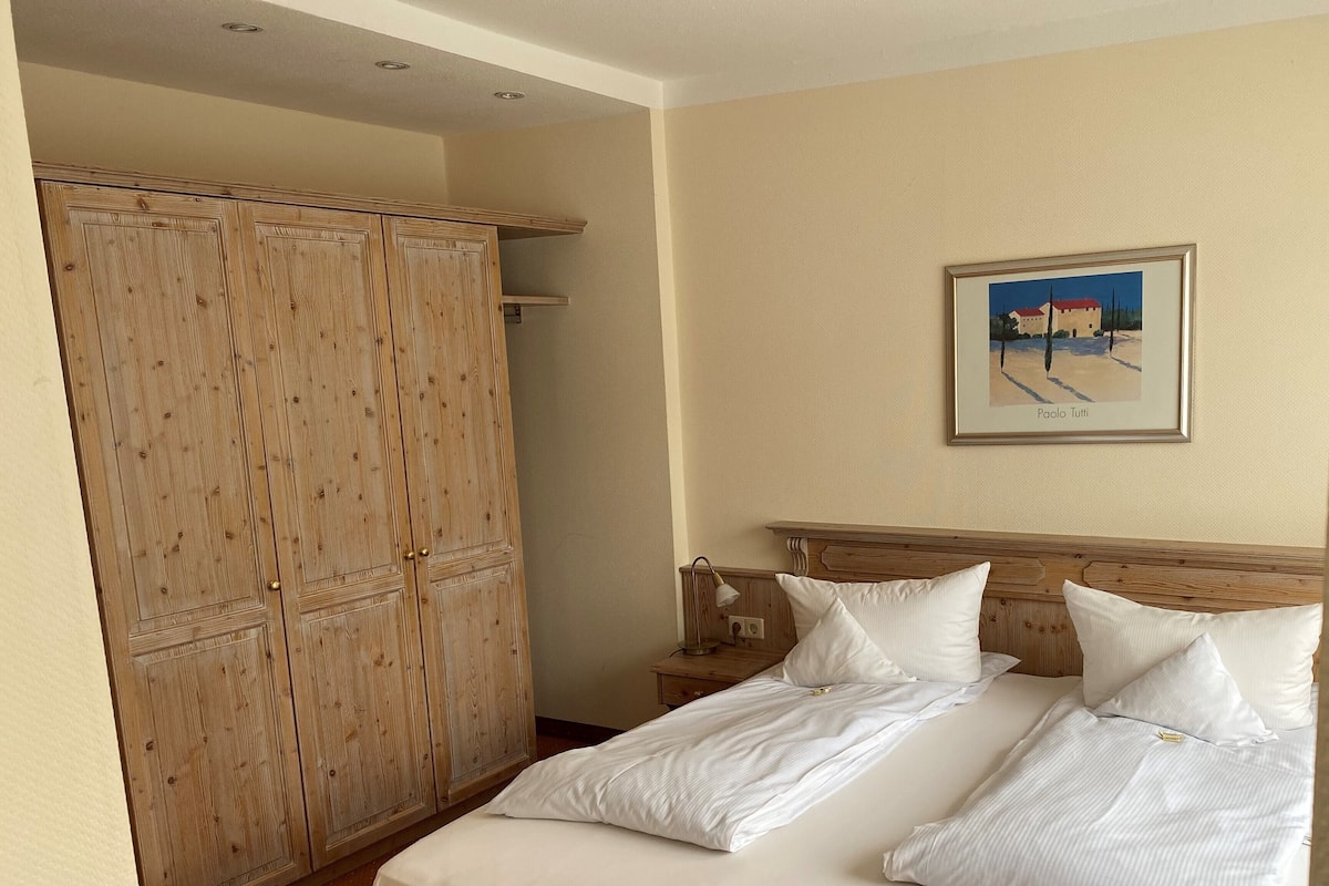 A hotel room with a large wooden wardrobe, a bed with white linens, and a framed painting on the wall.