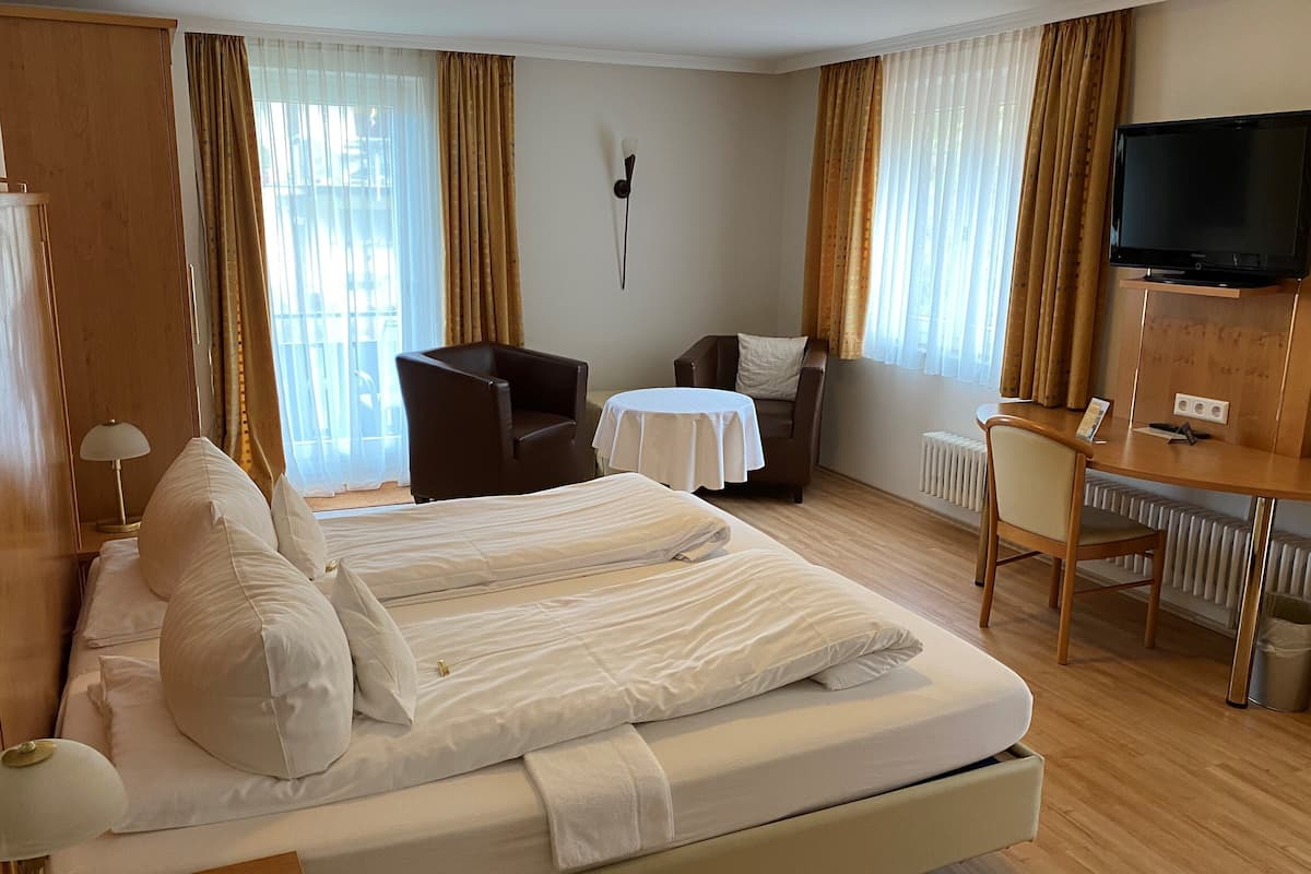 A hotel room with a large bed, two armchairs, a small table, a desk, and a TV.