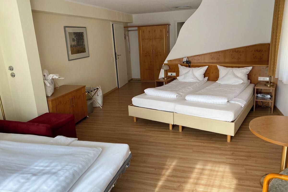 A hotel room with a large bed, a smaller bed, a wooden wardrobe, a desk with a lamp, and a small round table.