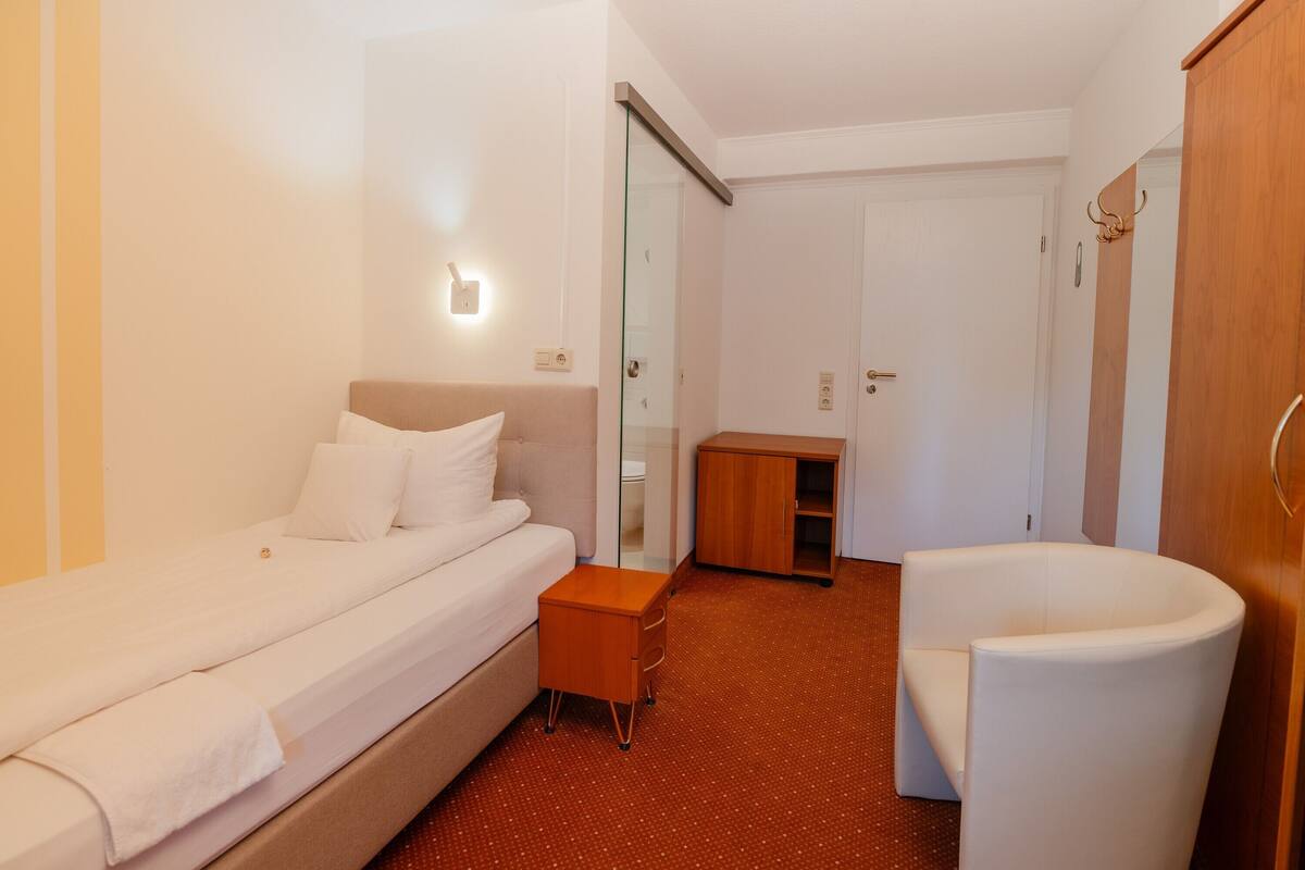 A hotel room with a bed, a small wooden cabinet, a white chair, and a glass door leading to a bathroom.