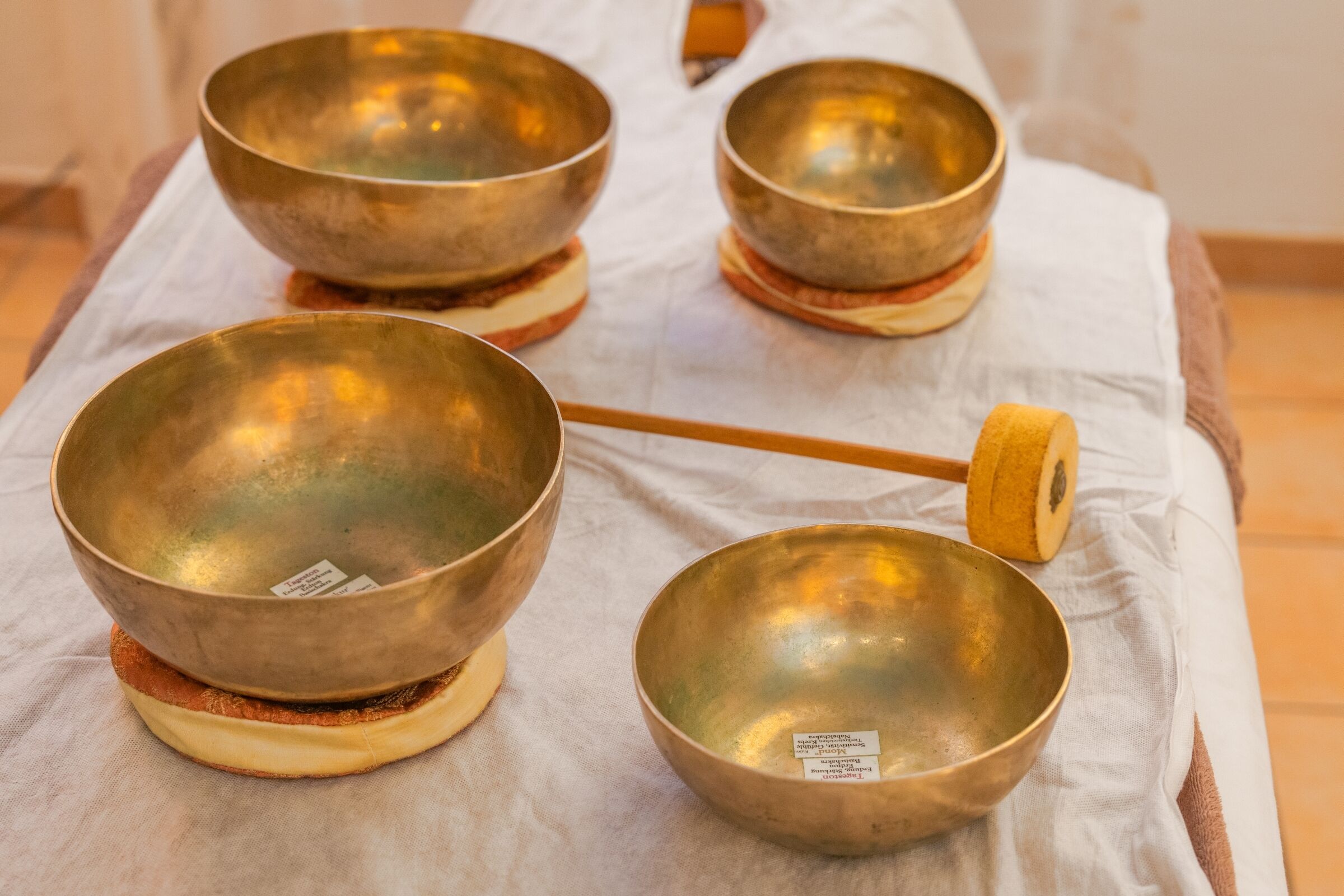 Four brass singing bowls on a white cloth with a wooden mallet.