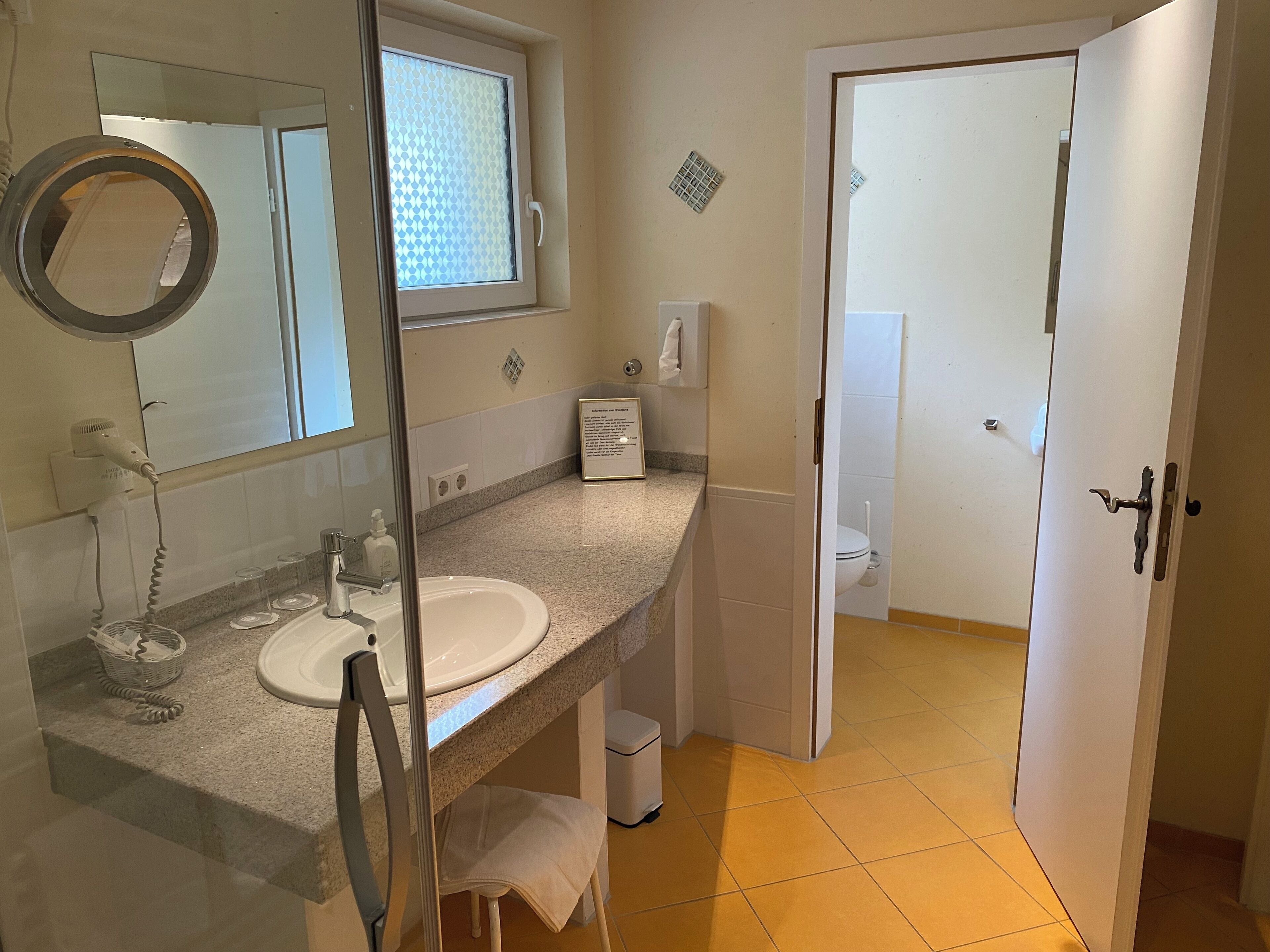 Ein Badezimmer mit Waschbecken, Spiegel und Fenster. Handtuchhalter und Mülleimer sind ebenfalls vorhanden.