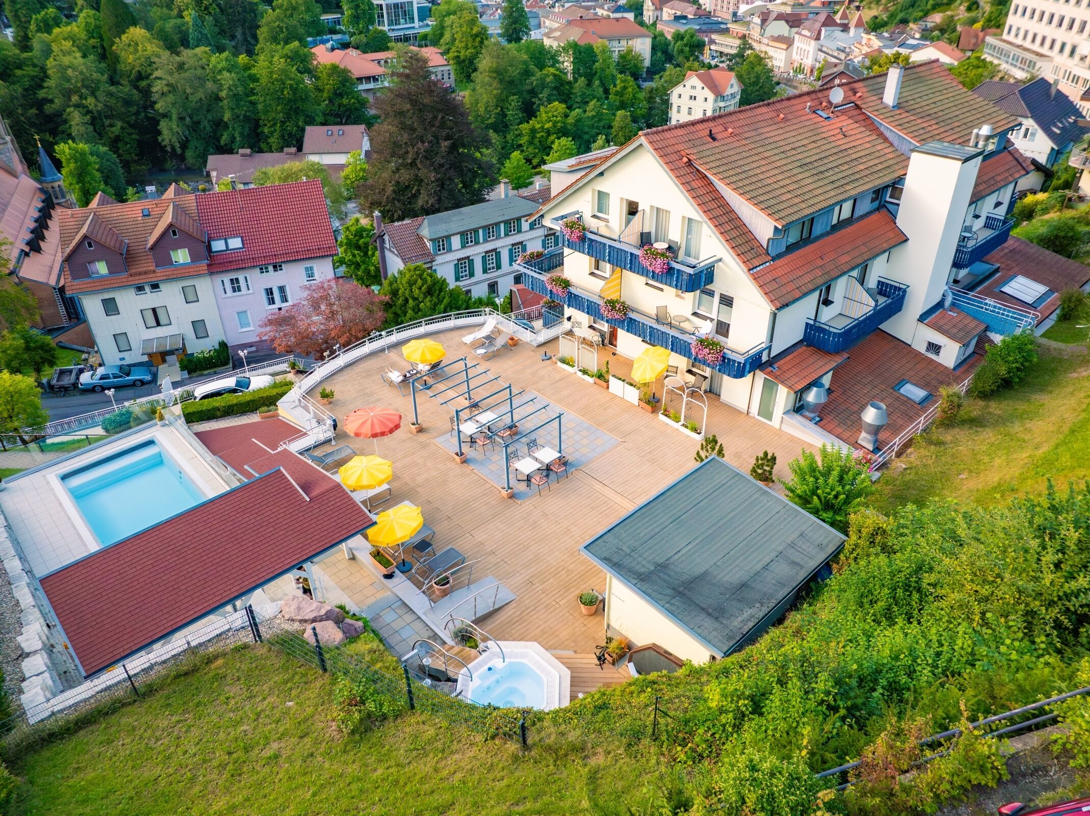 An aerial view of a residential area with a large house featuring a swimming pool, outdoor seating, and a balcony with hanging flower baskets.