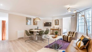A modern living room with a sofa, a dining table with chairs, a TV, and a large window with curtains. - Sweett | Madeleine I (Paris)