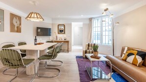 A modern living room with a sofa, a dining area with chairs, a TV, and a large window. - Sweett | Madeleine I (Paris)