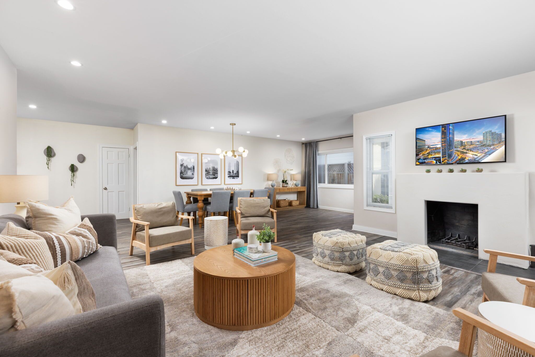 A modern living room with a sofa, ottoman, coffee table, and a fireplace. There is a dining area with chairs and a table, and a large TV mounted on the wall.