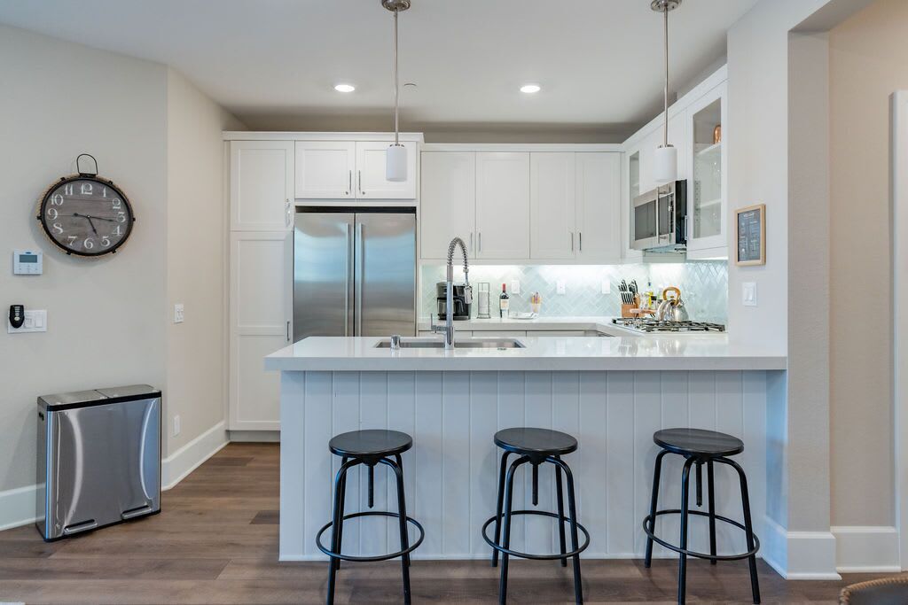 A modern kitchen with white cabinets, a stainless steel refrigerator, a sink, and three black bar stools.