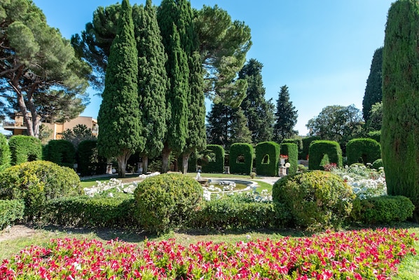 A well-manicured garden with various trees, a fountain, and flower beds.