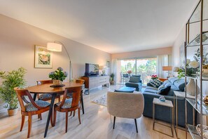 A modern living room with a dining area, a sofa, and a TV. There are plants and a window with curtains.