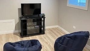 Una habitación con una televisión de pantalla plana sobre un soporte negro, dos sillones informales oscuros y un ventanal con persianas.