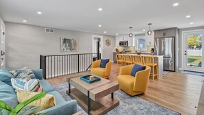 Un salon moderne avec un canapé, une table basse et des fauteuils jaunes. On aperçoit à l’arrière-plan un coin cuisine avec un réfrigérateur et un four à micro-ondes.