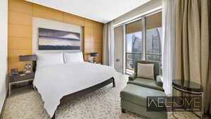 Una habitación de hotel moderna con una cama grande, un sillón verde, una mesita de noche con lámpara y un ventanal con vistas a la ciudad.