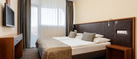 Una habitación de hotel con una cama grande, una mesita de noche, un televisor y un ventanal con cortinas.