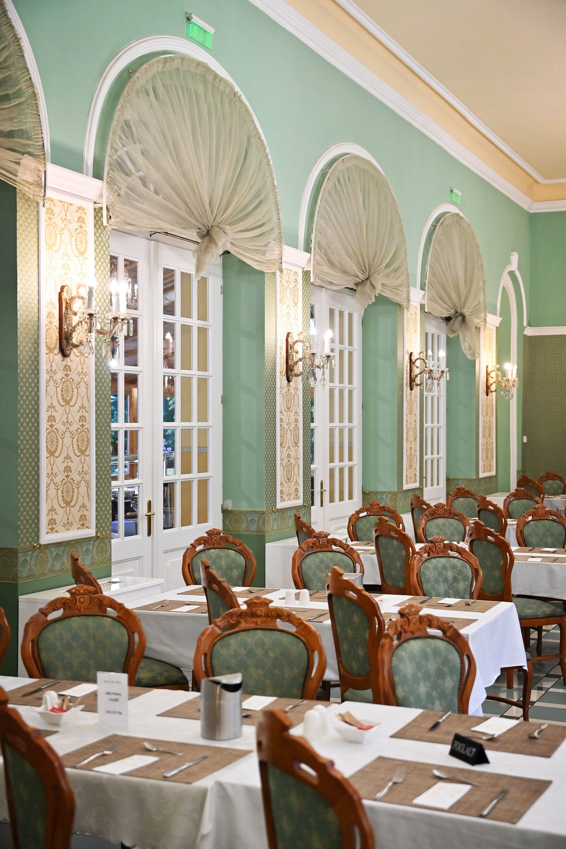 Un comedor con mesas dispuestas para el servicio, sillas con respaldos ornamentados y apliques decorativos en la pared.