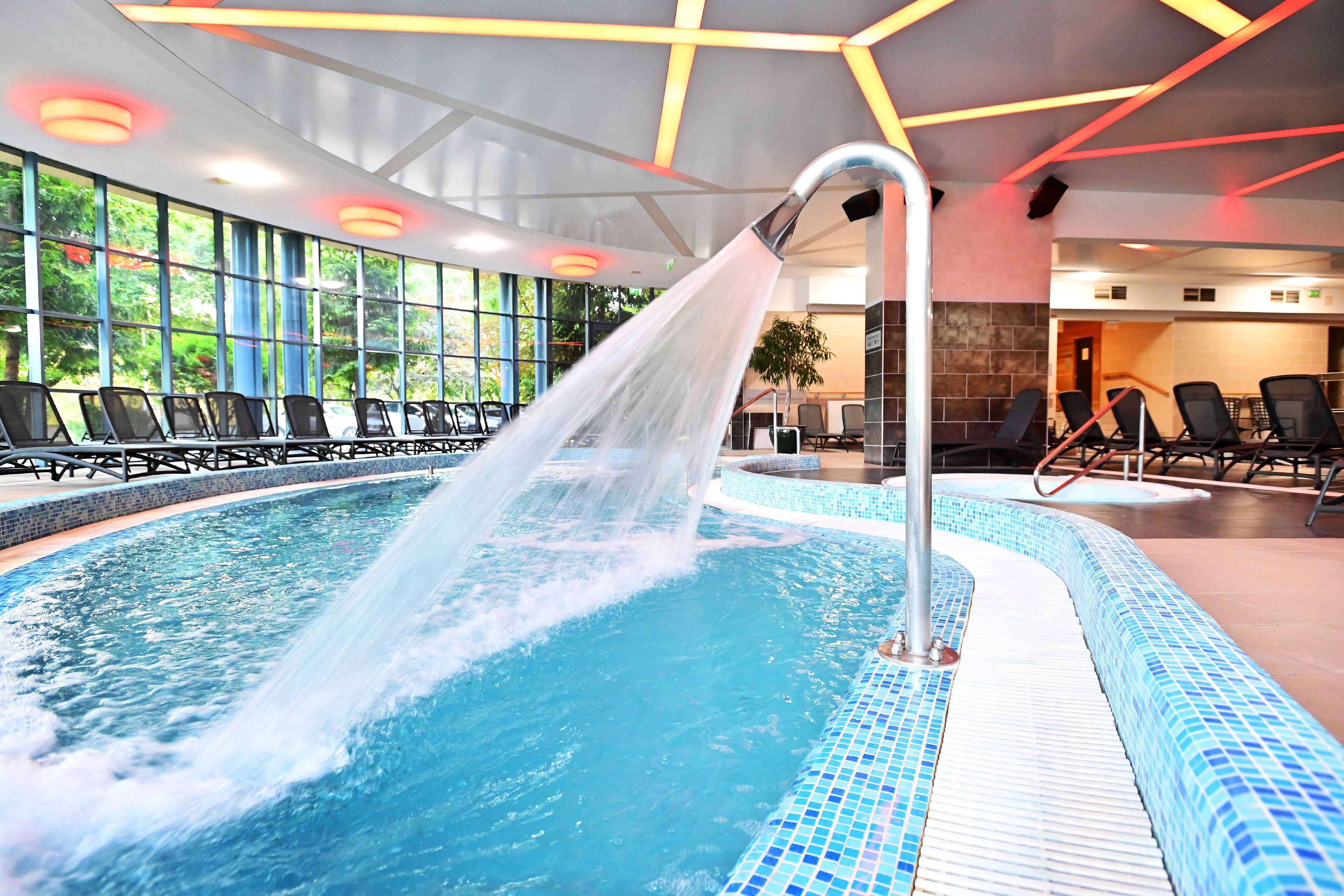 Une piscine intérieure agrémentée d’un élément décoratif aquatique, entourée de chaises longues et dotée de grandes fenêtres.