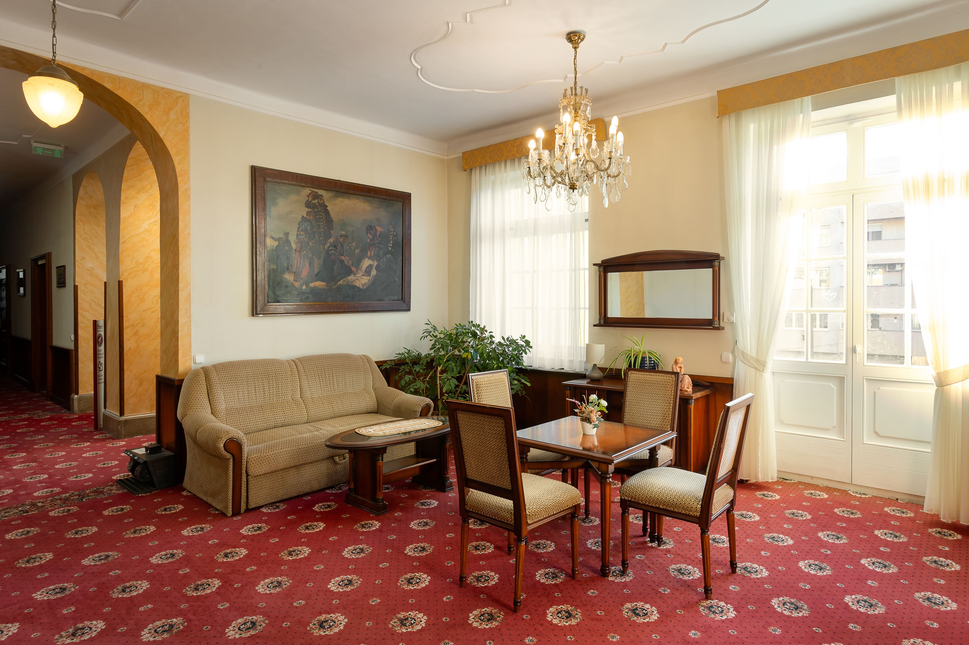 A room with a red patterned carpet, a sofa, a dining table with chairs, a chandelier, a painting on the wall, and a large window with curtains.