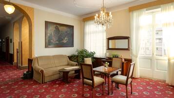 A room with a red patterned carpet, a sofa, a dining table with chairs, a chandelier, a painting on the wall, and a large window with curtains.
