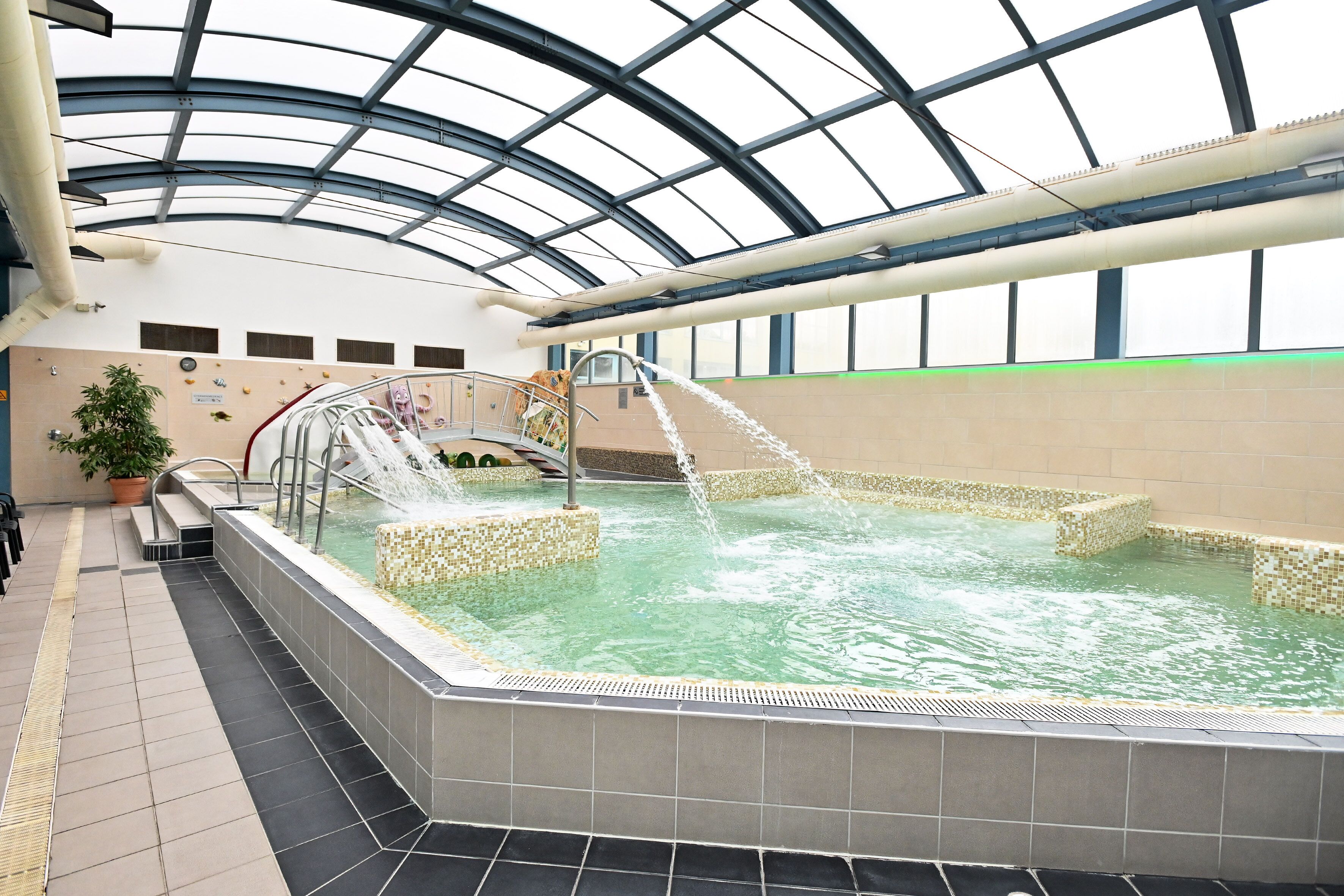 An indoor swimming pool with a glass ceiling, tiled walls, and a water fountain.