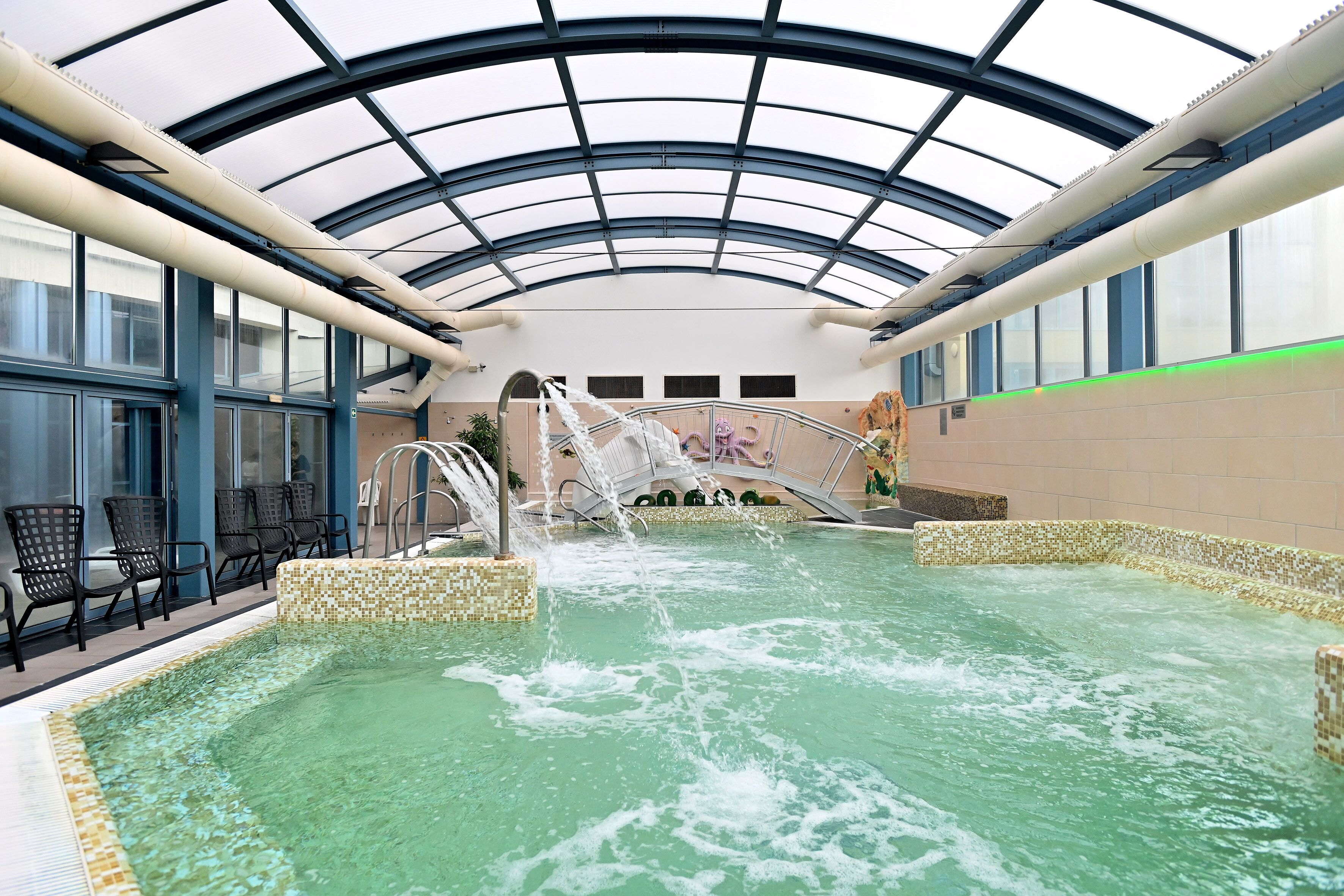 Una piscina interior con techo de cristal, tobogán acuático y zona de estar con sillones.
