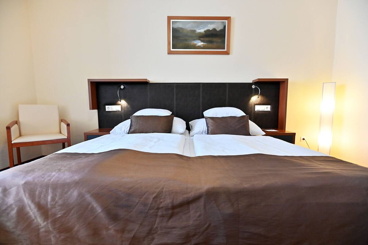 A double bed with a brown bedspread, two bedside tables with lamps, a chair, and a framed picture on the wall.