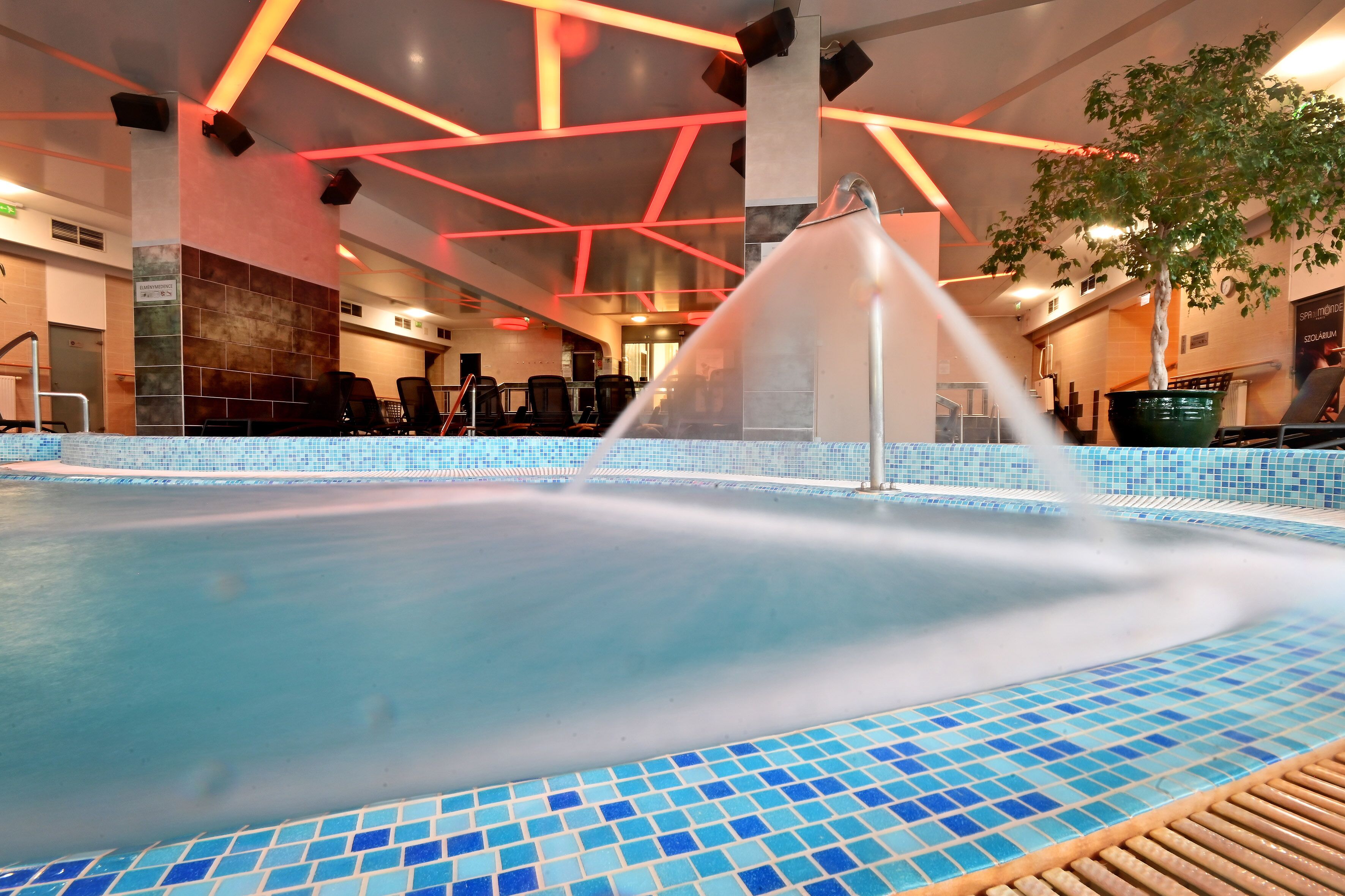 An indoor swimming pool with a water fountain, tiled walls, and ceiling lights.