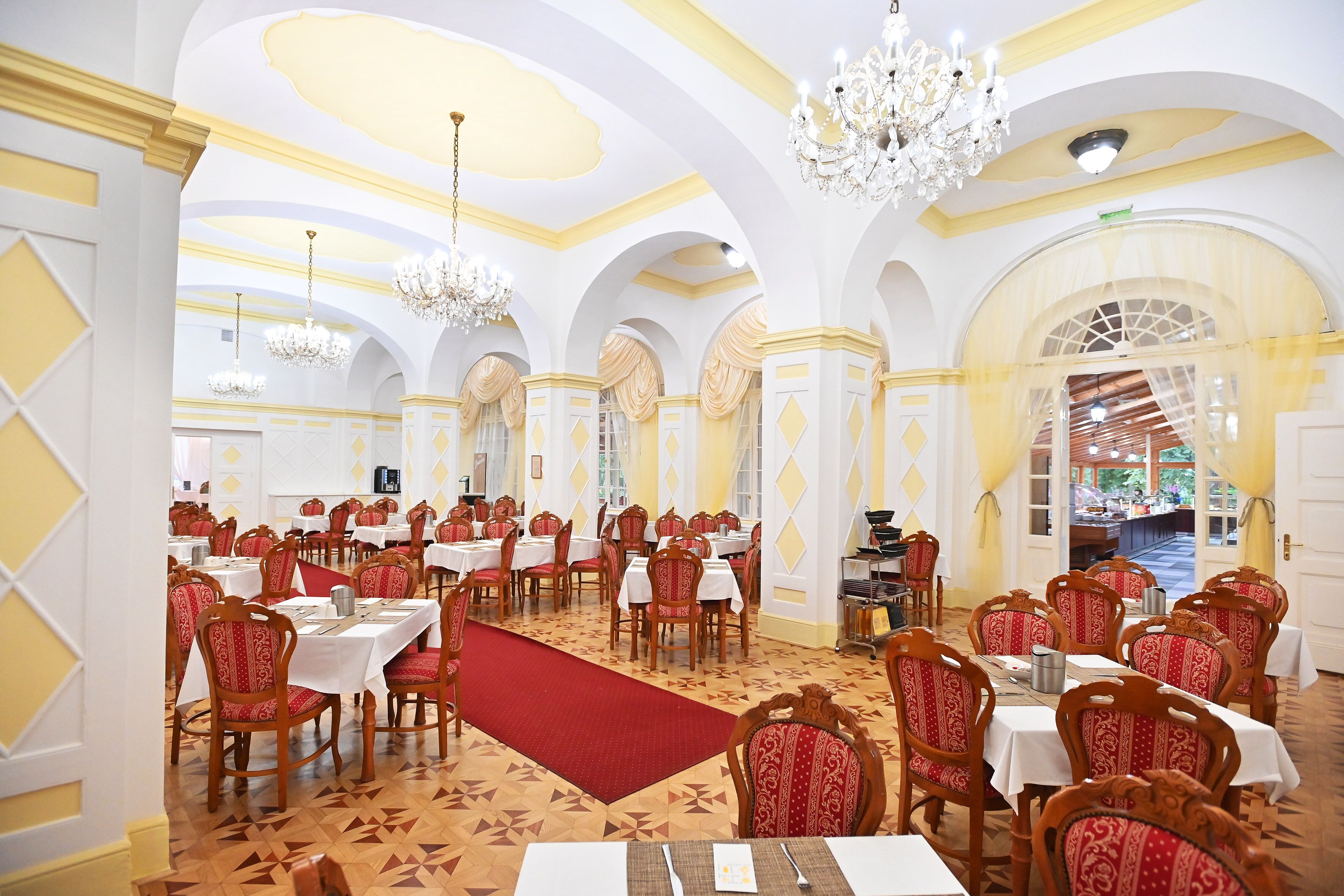 A spacious dining area with multiple tables and chairs, chandeliers, and a red carpet.