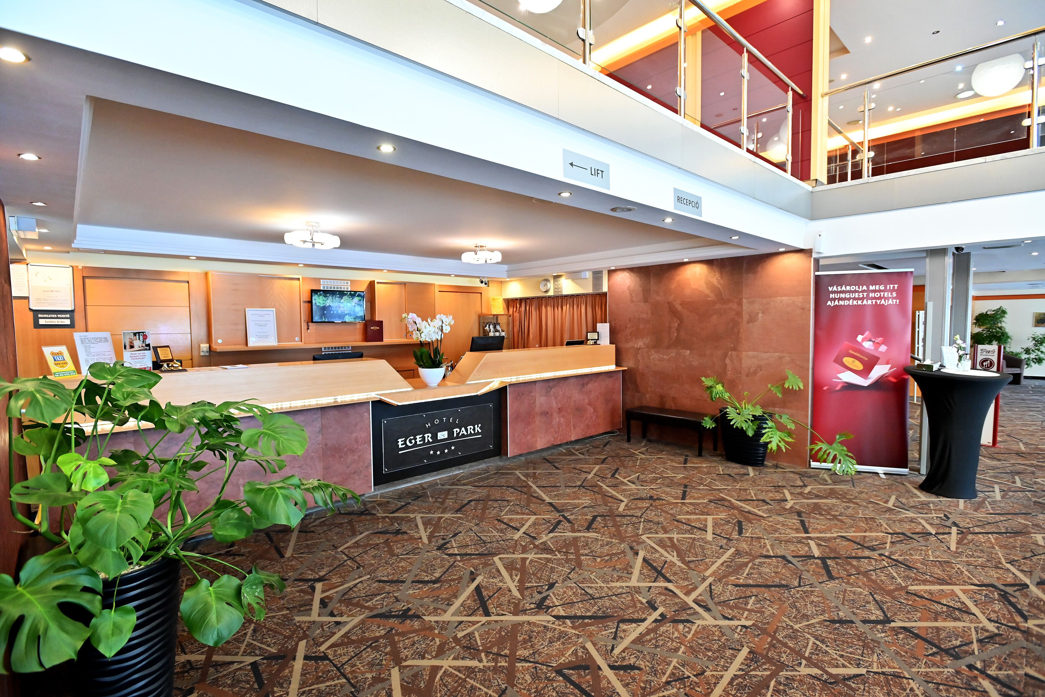 A hotel reception area with a modern design, including a reception desk, a television, and a sign indicating the direction to the lift and reception.