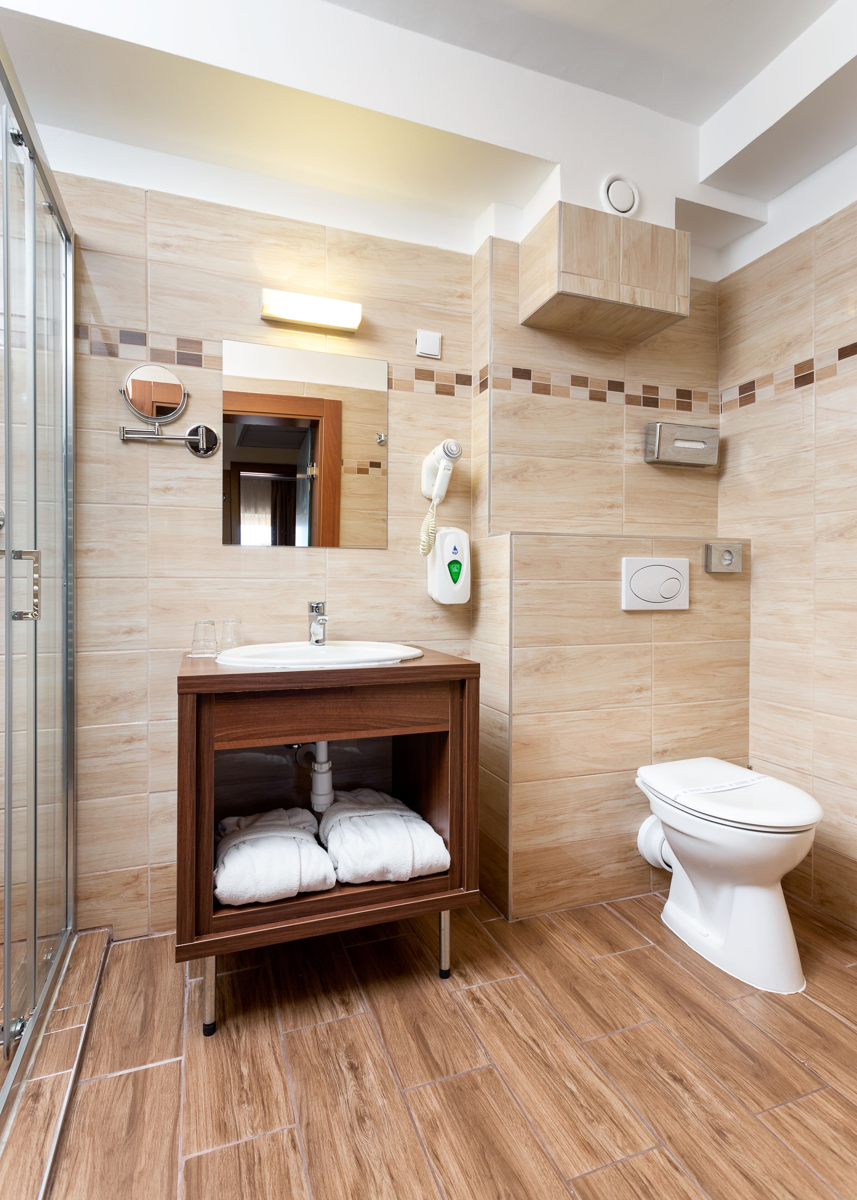 A modern bathroom with a wooden vanity, a toilet, a glass shower enclosure, and a mirror with a built-in light.
